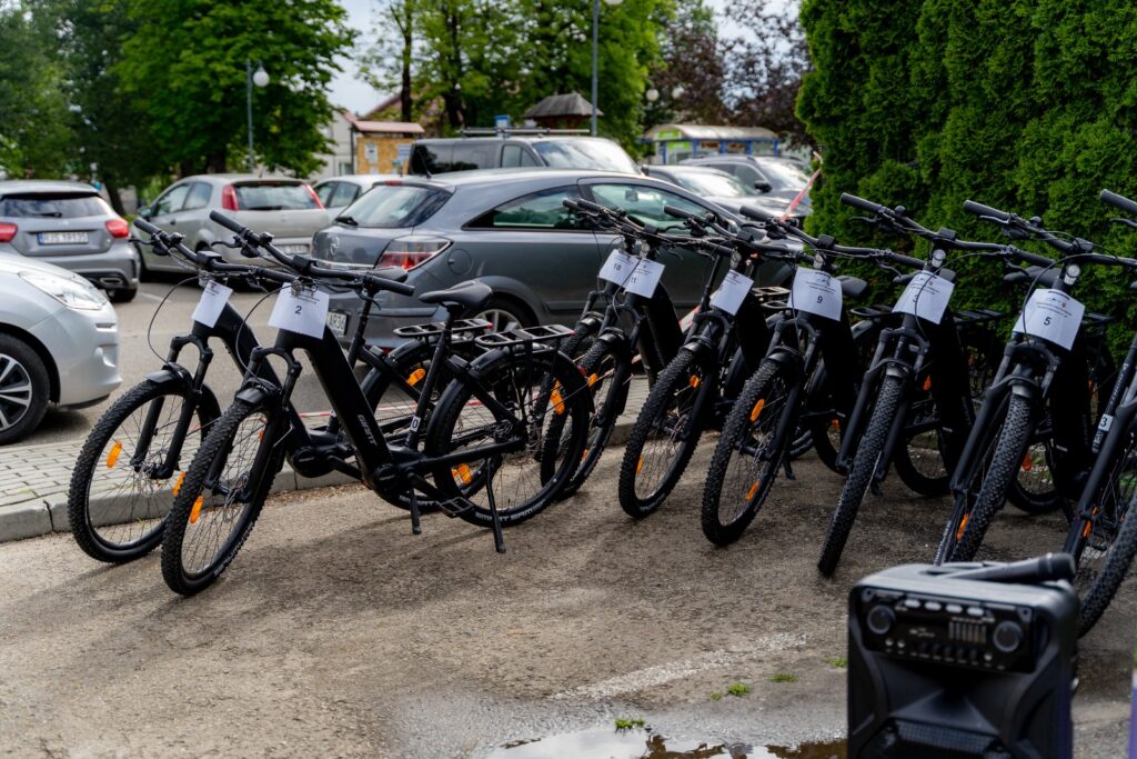 Rowery elektryczne zaparkowane przy krawężniku na parkingu, w tle zaparkowane samochody osobowe.