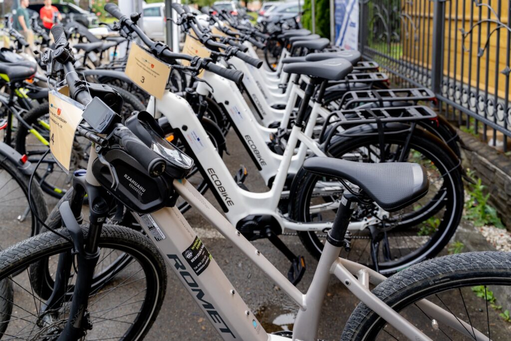 Rząd białych rowerów elektrycznych marek Ecobike i Romet z zawieszonymi tabliczkami startowymi.