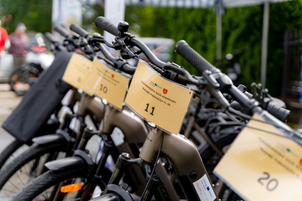 Rad zaparkovaných elektrických bicyklov so zavesenými štartovými číslami, v popredí číslo 11 s nápisom „Cezhraničný cyklistický rely po trasách pohraničia PL-SK“.