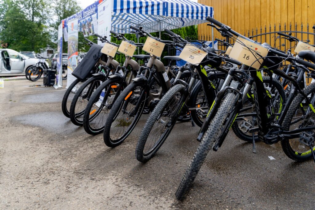 Rząd rowerów elektrycznych z tabliczkami startowymi ustawiony przed namiotem z napisem „Gmina Osiek Jasielski”, w tle rollupy informacyjne i zaparkowane samochody.