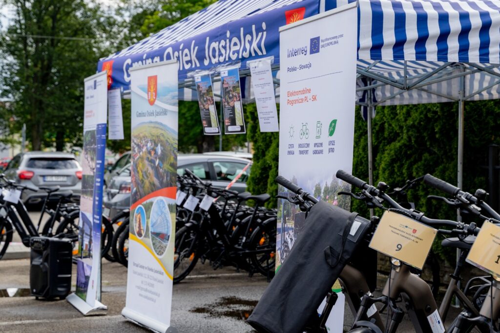 Rollupy reklamowe „Gmina Osiek Jasielski” i „Interreg – Elektromobilne Pogranicze PL-SK” ustawione przy namiocie, obok zaparkowane rowery elektryczne z tabliczką startową numer 9.