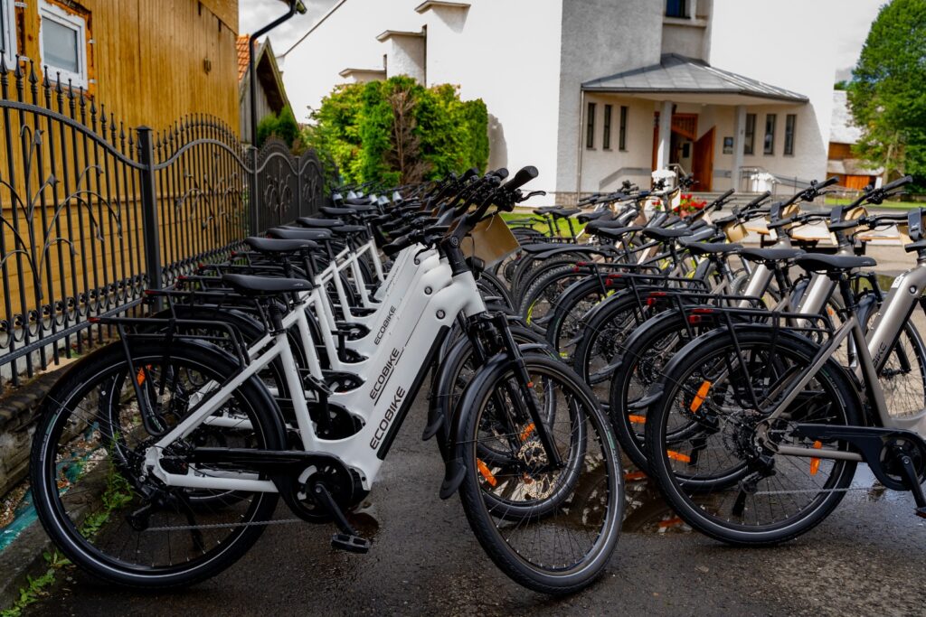 Rząd białych rowerów elektrycznych marki Ecobike zaparkowanych wzdłuż kutego ogrodzenia, w tle budynek kościoła.