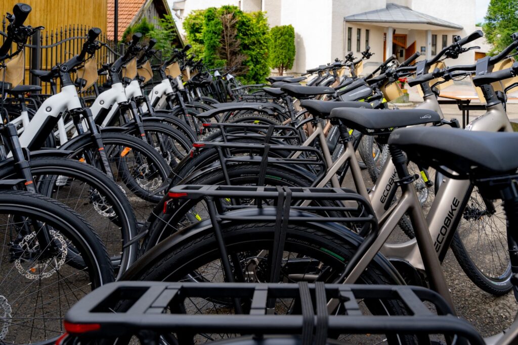 Rząd bagażników na tylnych kołach gęsto zaparkowanych rowerów elektrycznych, w tle budynek kościoła.