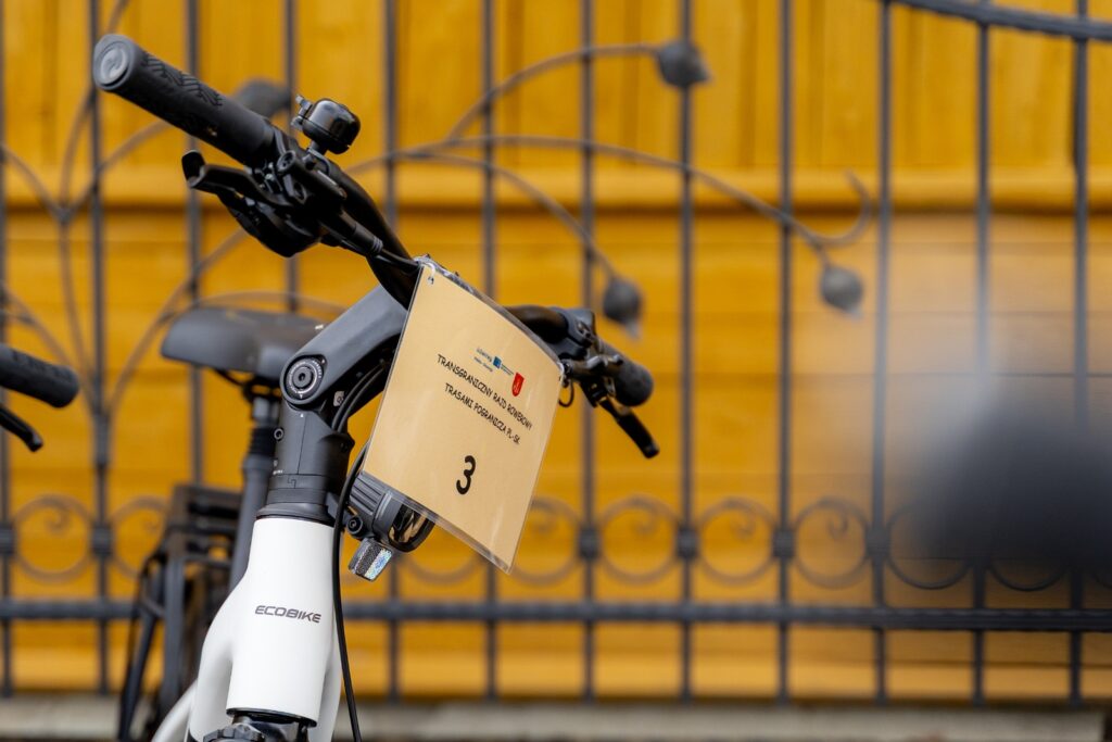 Biały rower elektryczny marki Ecobike z zawieszoną tabliczką startową numer 3, w tle kuta metalowa brama i żółta drewniana ściana.