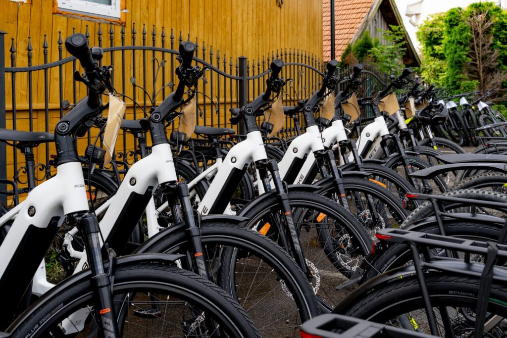 Rząd białych rowerów elektrycznych marek Ecobike i Romet oparty o kute ogrodzenie na tle żółtej ściany.