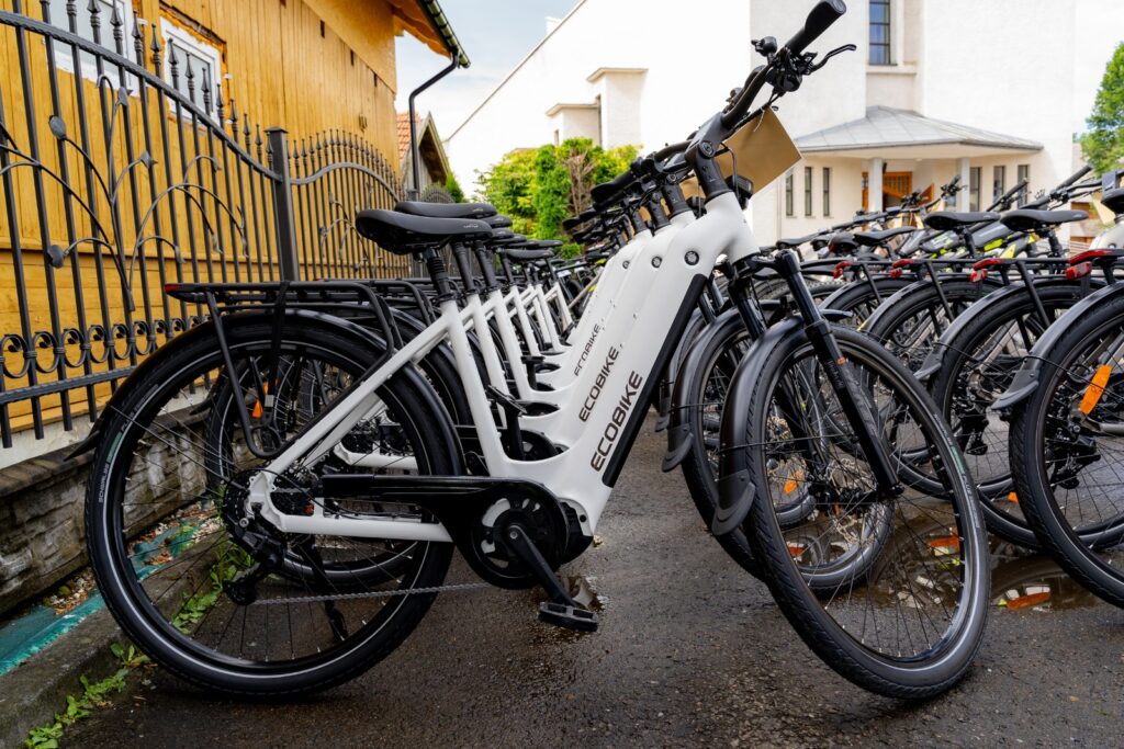 Biały rower elektryczny marki Ecobike z zawieszoną tabliczką startową na pierwszym planie, w tle rząd czarnych rowerów i budynek kościoła.