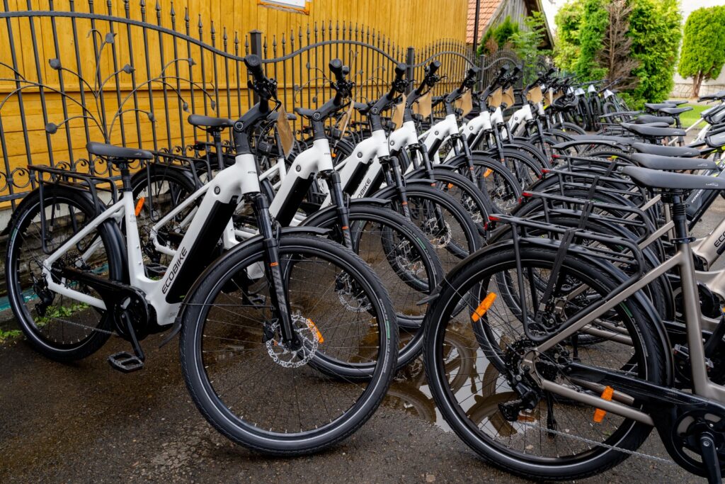 Rząd białych rowerów elektrycznych marki Ecobike ustawiony wzdłuż kutego ogrodzenia na mokrej nawierzchni.