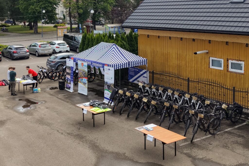Widok z góry na namiot i stoły z materiałami organizacyjnymi oraz rowery elektryczne ustawione rzędem wzdłuż żółtego budynku, w tle zaparkowane samochody.