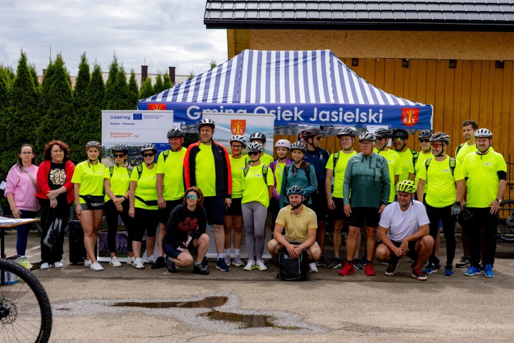 Grupa rowerzystów w żółtych koszulkach odblaskowych i kaskach pozująca do zdjęcia przed namiotem z napisem „Gmina Osiek Jasielski”.