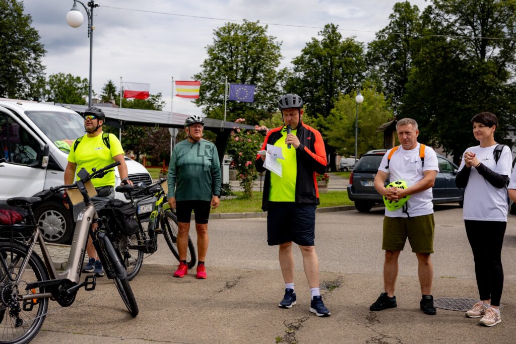 Grupa osób stojących na placu, mężczyzna przemawia do mikrofonu, w tle flagi