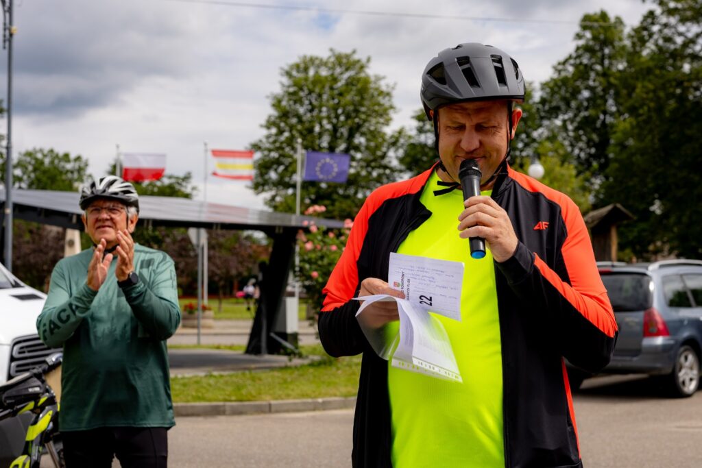 Mężczyzna w kasku rowerowym przemawia do mikrofonu, trzymając kartkę , obok oklaskujący go mężczyzna, w tle flagi.