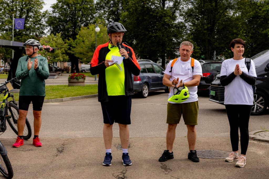 Czworo uczestników rajdu stojących na placu, mężczyzna w kasku przemawia do mikrofonu, pozostali klaszczą, w tle zaparkowane samochody.