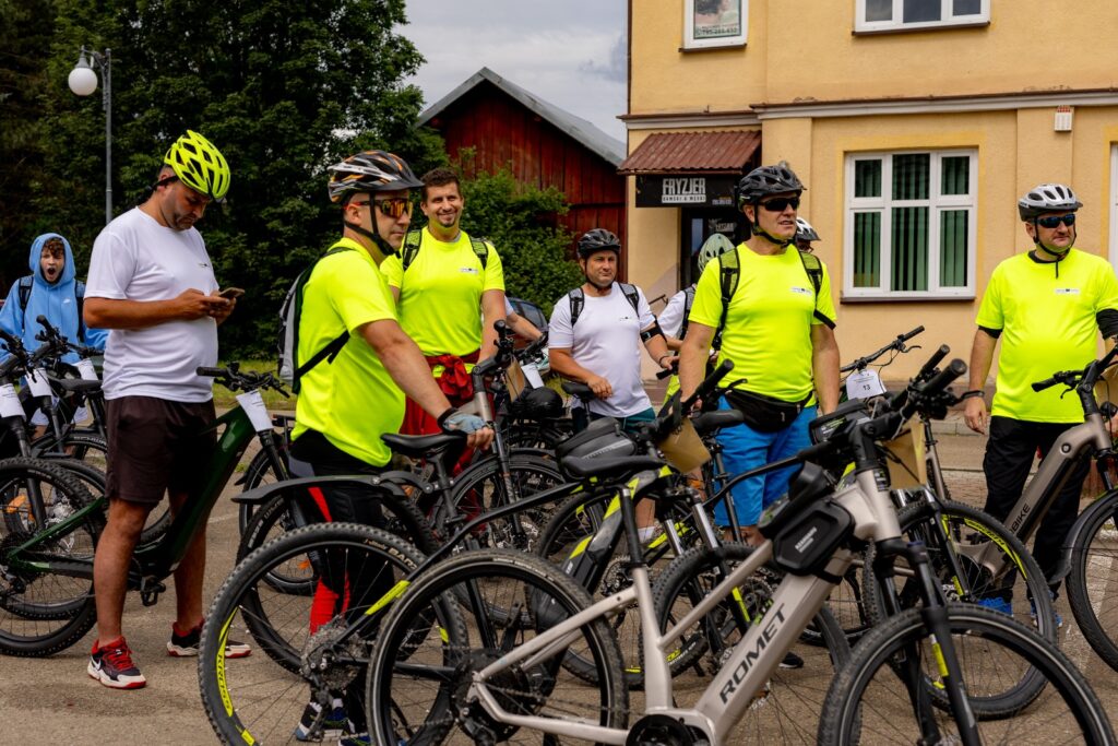 Grupa rowerzystów w kaskach i koszulkach odblaskowych stojąca przy rowerach, w tle żółty budynek z szyldem zakładu fryzjerskiego.