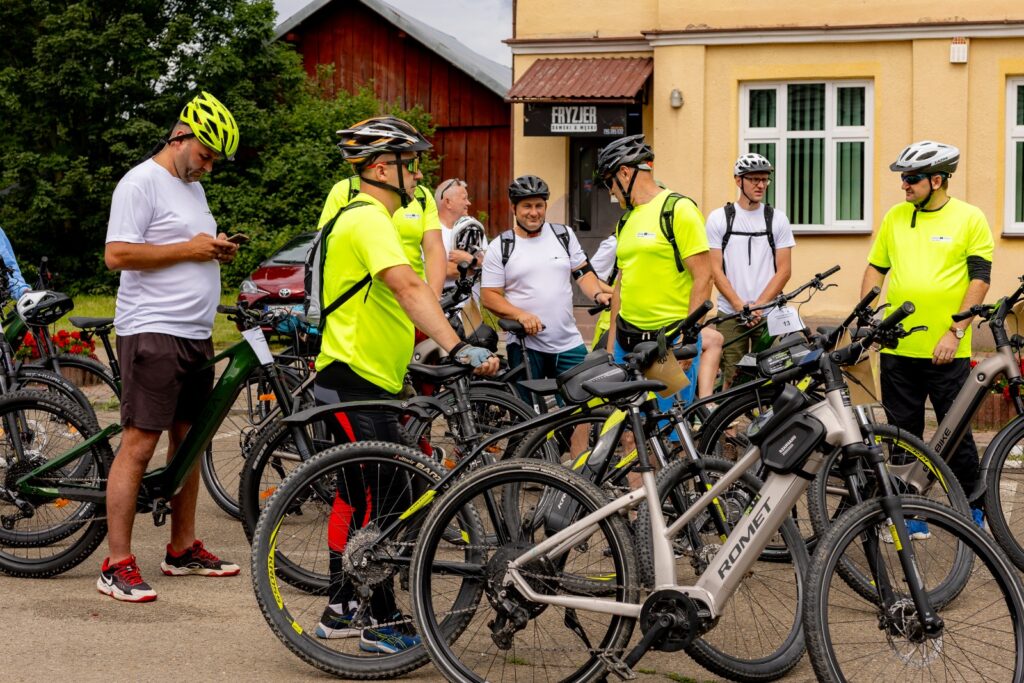 Liczniejsza grupa rowerzystów w kaskach zebrana przy rowerach na placu przed budynkiem z szyldem zakładu fryzjerskiego.