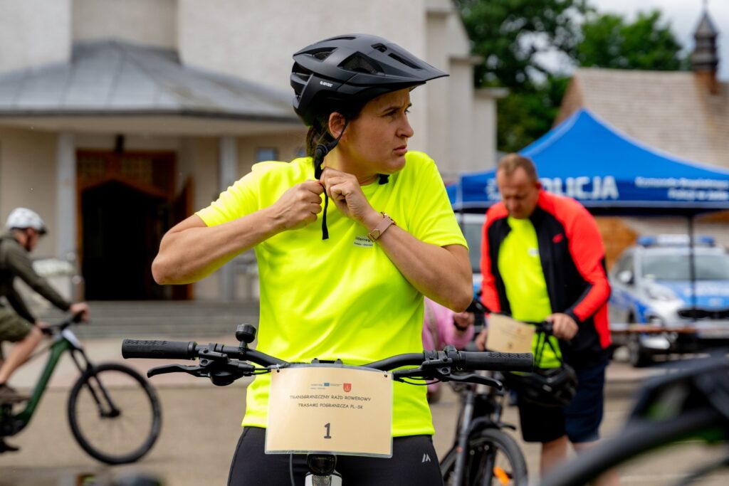 Kobieta zapinająca kask rowerowy przy rowerze z tabliczką startową, w tle niebieski namiot policyjny i radiowóz.