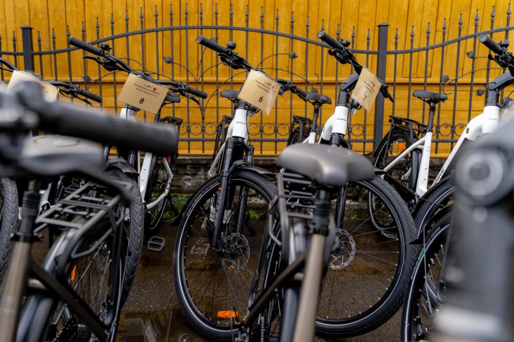 Rząd rowerów elektrycznych z numerami startowymi 6, 7 i 8 oparty o kutą bramę na tle żółtej ściany.