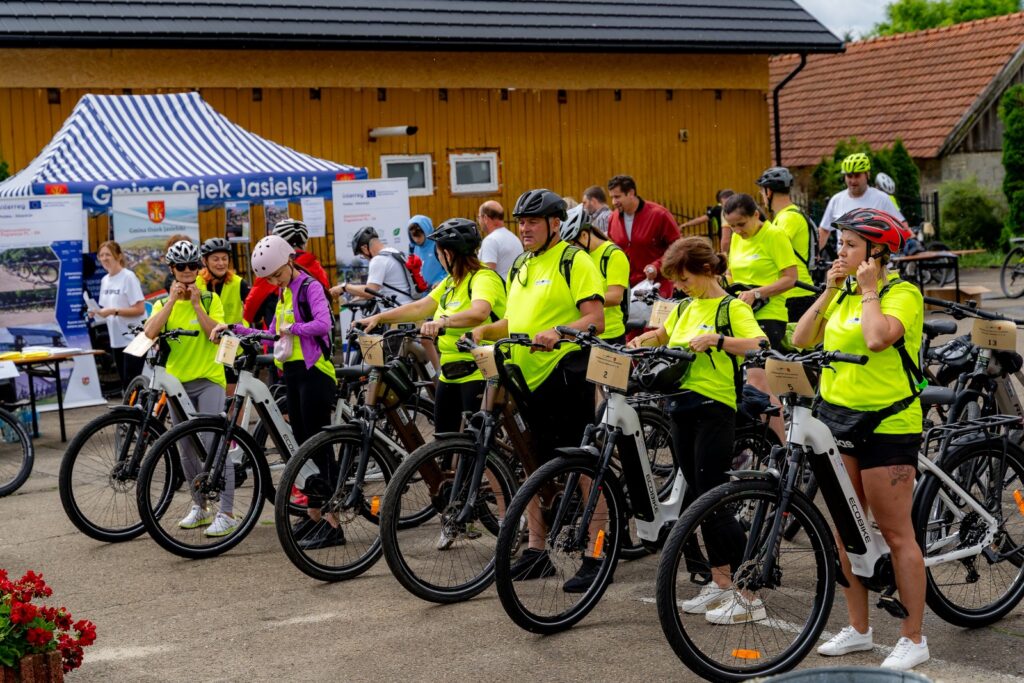 Grupa rowerzystów w żółtych koszulkach odblaskowych zakładających kaski przy rowerach, w tle namiot z napisem „Gmina Osiek Jasielski”.