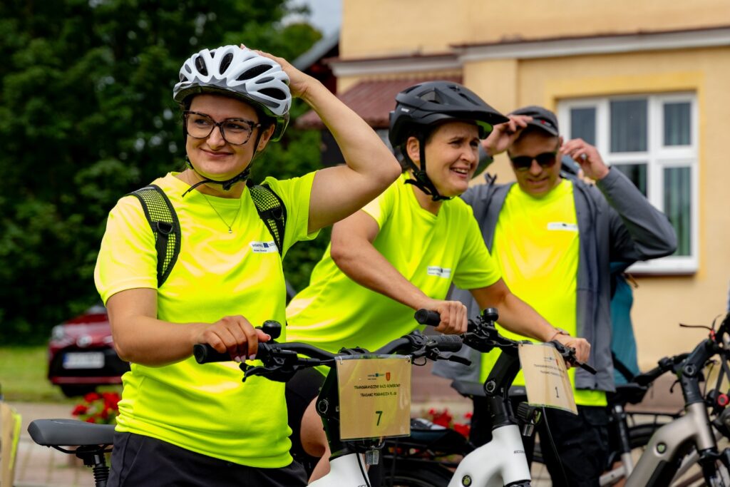 Troje rowerzystów w żółtych koszulkach odblaskowych zakładających kaski, przy rowerach z tabliczkami startowymi
