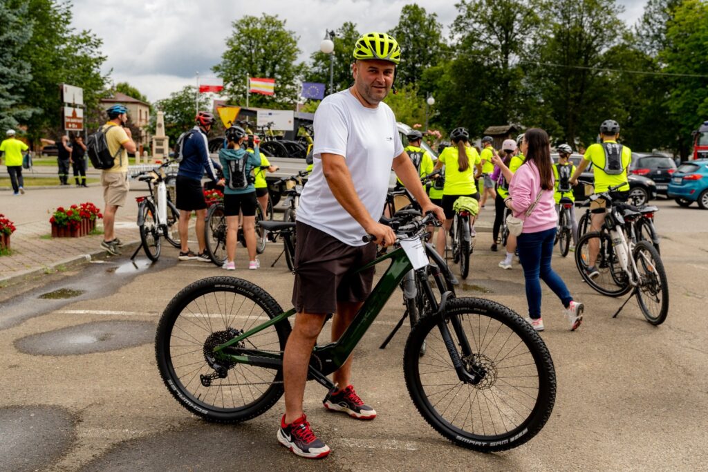 Mężczyzna w żółtym kasku rowerowym stojący przy zielonym rowerze górskim, w tle flagi i grupa rowerzystów.