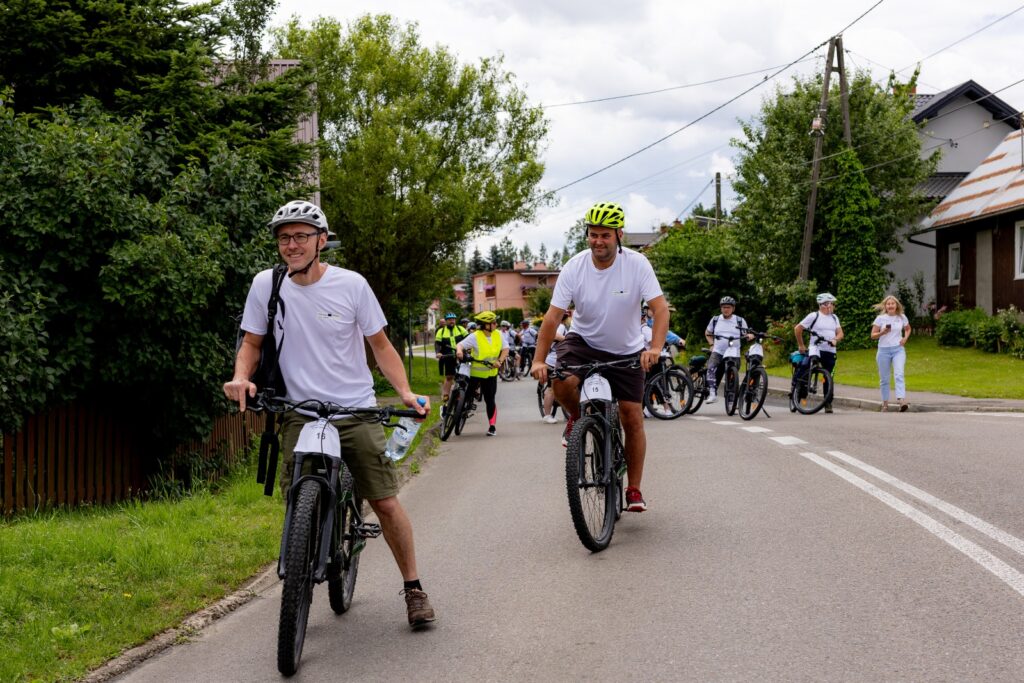 Dwaj rowerzyści z tabliczkami startowymi, jadący ulicą, w tle grupa pozostałych uczestników rajdu.