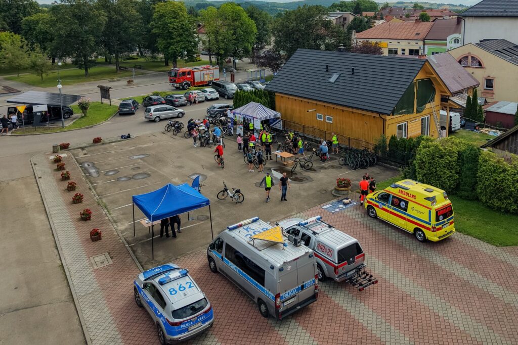 Widok z lotu ptaka na plac z namiotem i zaparkowanymi pojazdami służb: policji, straży pożarnej i pogotowia ratunkowego, obok żółty budynek.