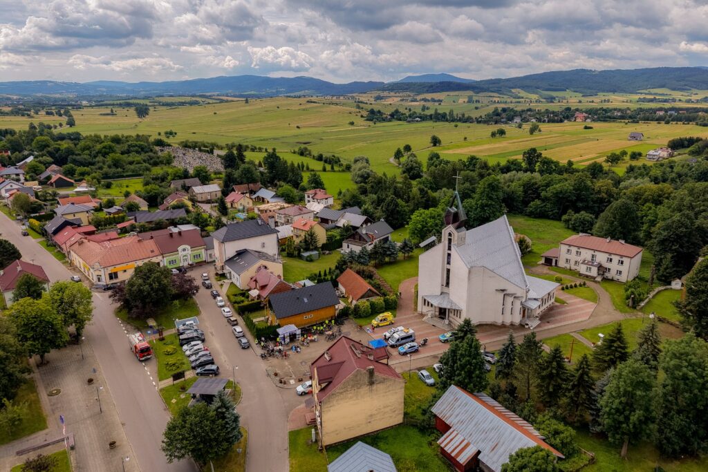 Widok z lotu ptaka na wieś z kościołem, zabudową i polami w tle gór, na placu przed kościołem widoczna grupa uczestników rajdu.