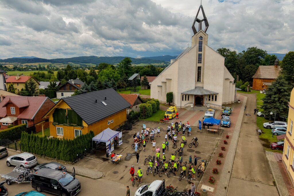 Widok z lotu ptaka na kościół i plac z grupą rowerzystów, pojazdami policji i karetką pogotowia, w tle góry.
