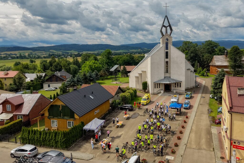 Widok z lotu ptaka na kościół i plac z grupą rowerzystów, karetką pogotowia oraz budynkiem poczty w tle.