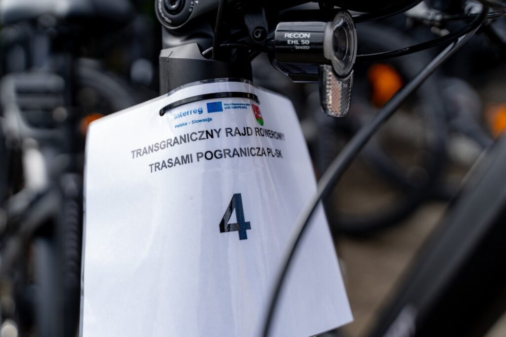 Zbliżenie tabliczki startowej z numerem 4 i logo Interreg zawieszonej na kierownicy roweru.