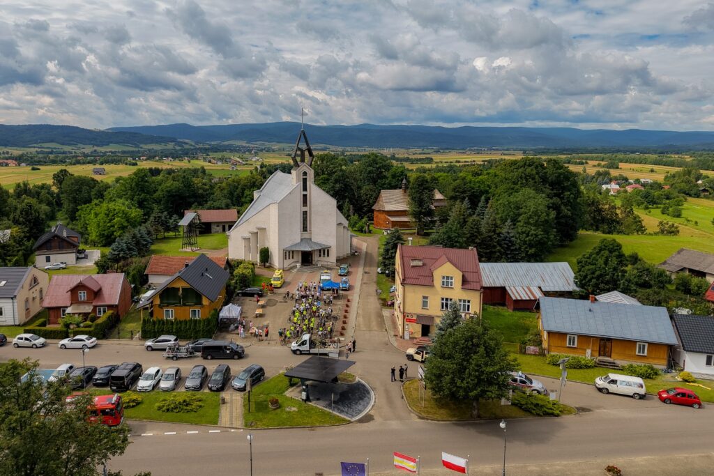 Widok z lotu ptaka na wieś z kościołem, zaparkowanymi samochodami i grupą rowerzystów na placu, w tle łańcuch górski i flagi.