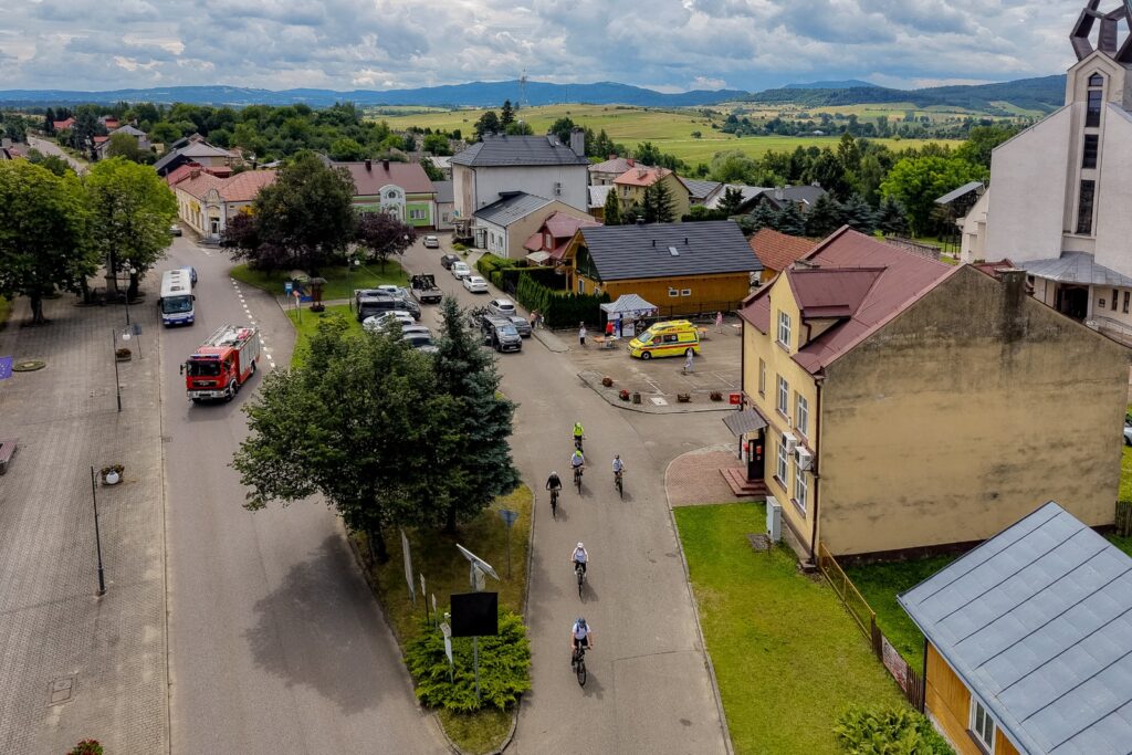 Widok z lotu ptaka na ulicę, którą jedzie kilku rowerzystów, w tle wóz strażacki, karetka pogotowia i zabudowa miejscowości.