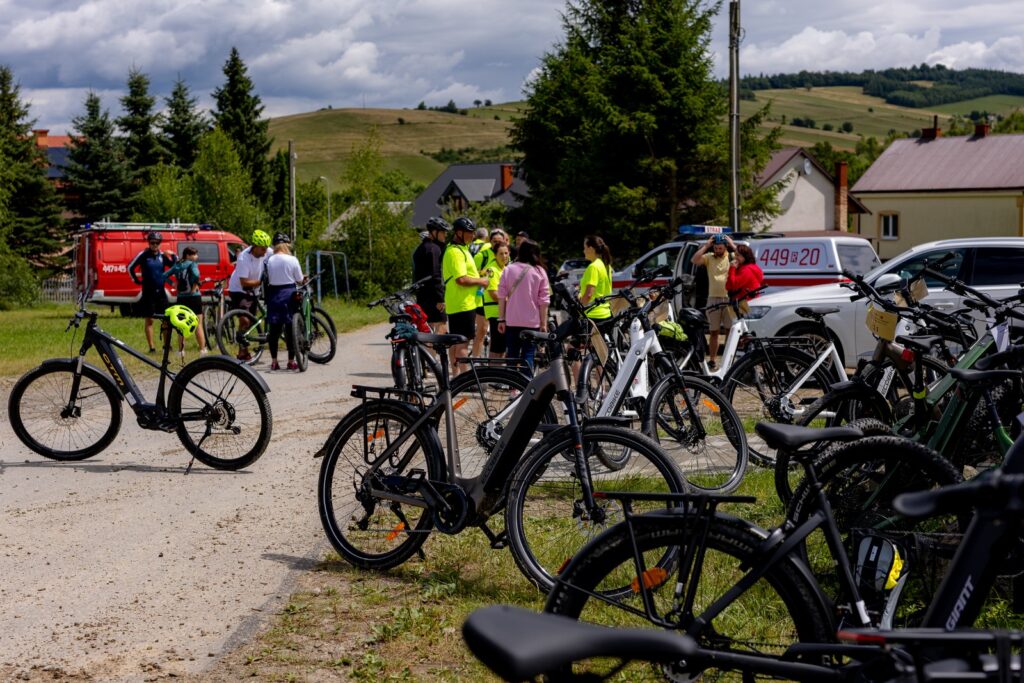 Grupa rowerzystów i zaparkowane rowery na drodze polnej, w tle wóz strażacki, radiowóz i pojazd pogotowia.