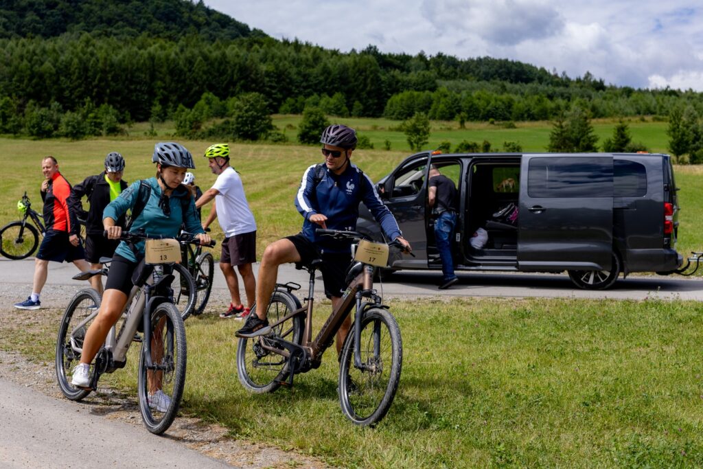Dwoje rowerzystów z tabliczkami startowymi, wsiadających na rowery, w tle otwarty czarny samochód dostawczy i łąka.