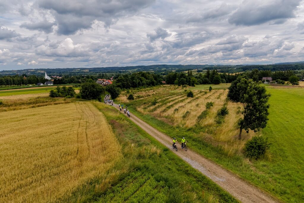Widok z lotu ptaka na grupę rowerzystów jadących polną drogą wśród pól zbóż, w tle wieża kościoła.