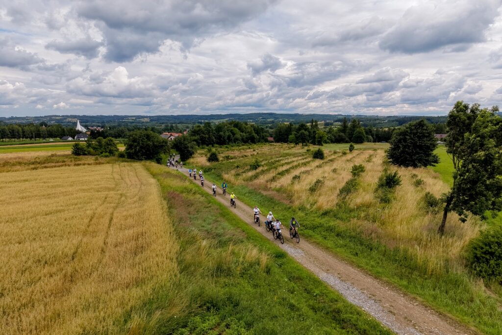 Widok z lotu ptaka na kolumnę rowerzystów jadących żwirową drogą pomiędzy polami, w tle zabudowania wsi.