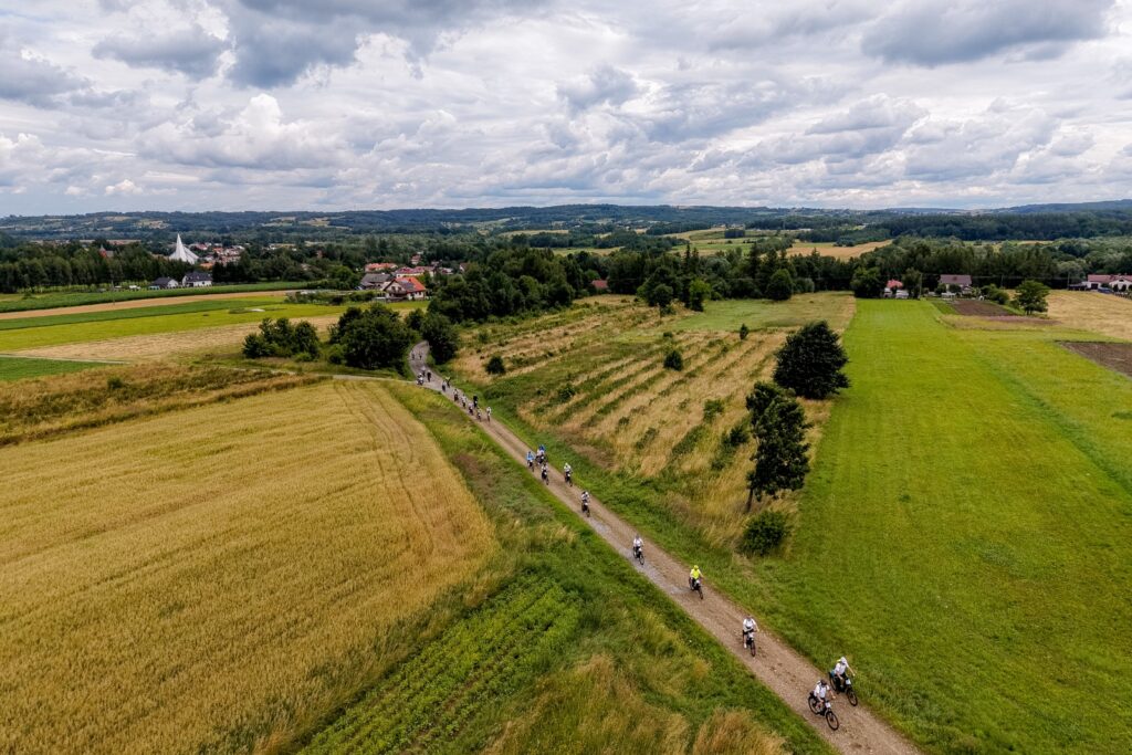 Widok z lotu ptaka na rowerzystów jadących drogą polną wśród pól uprawnych, w tle wieś i wzgórza.