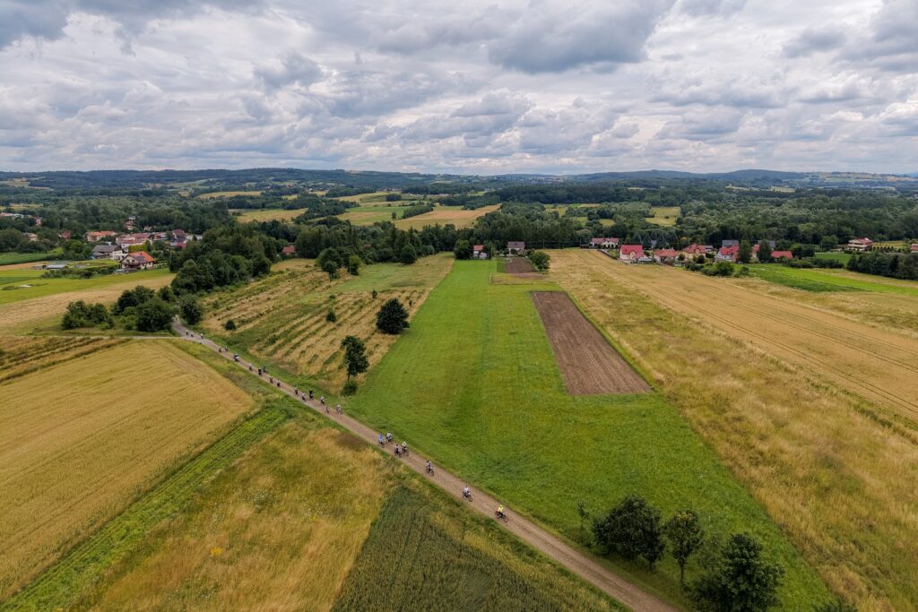 Widok z lotu ptaka na długą kolumnę rowerzystów jadących polną drogą przez pola, w tle zabudowa wsi po obu stronach.