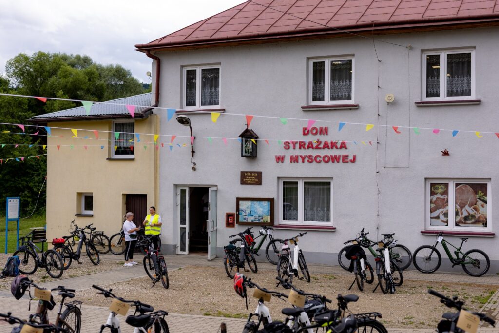 Budynek „Dom Strażaka w Myscowej” udekorowany kolorowymi flagami, przed nim zaparkowane rowery elektryczne.