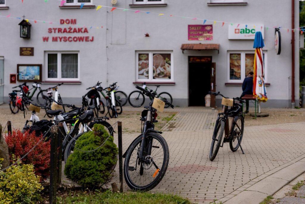 Elewacja Domu Strażaka w Myscowej ze sklepem spożywczo-przemysłowym, przed budynkiem zaparkowane rowery z tabliczkami startowymi.