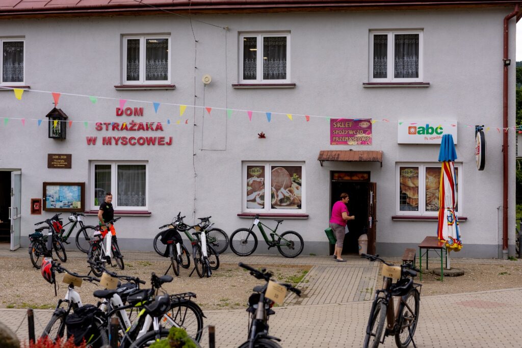 Budynek Domu Strażaka w Myscowej ze sklepem, kobieta wchodząca do sklepu, przed budynkiem zaparkowane rowery.