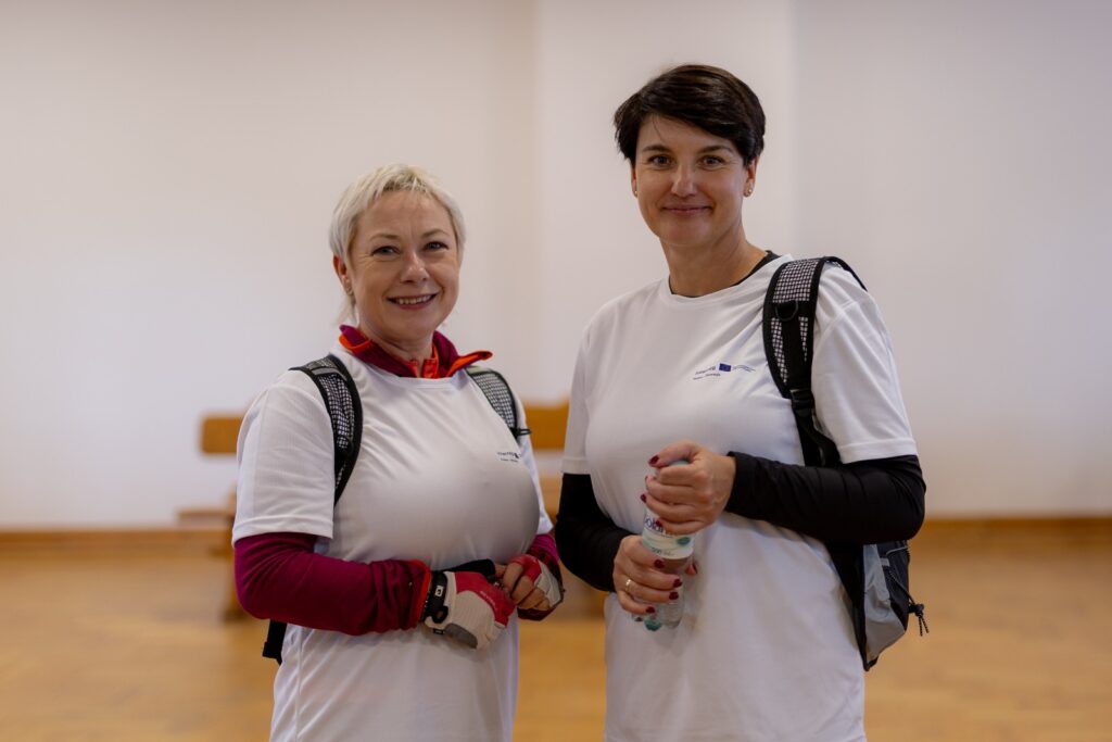 Dwie uśmiechnięte kobiety w białych koszulkach z plecakami pozujące do zdjęcia we wnętrzu sali.