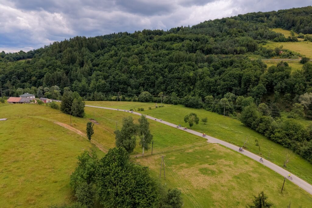 Widok z lotu ptaka na drogę wijącą się przez zalesione wzgórza, którą jedzie grupa rowerzystów.