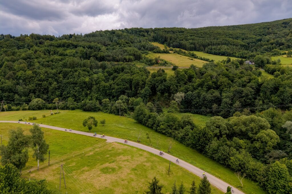 Widok z lotu ptaka na krętą drogę w dolinie otoczonej lasem, którą jedzie kilku rowerzystów.