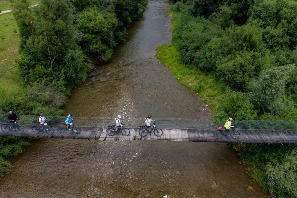 Widok z lotu ptaka na rowerzystów przejeżdżających przez wiszący kładkowy most nad rzeką, otoczony zielenią.