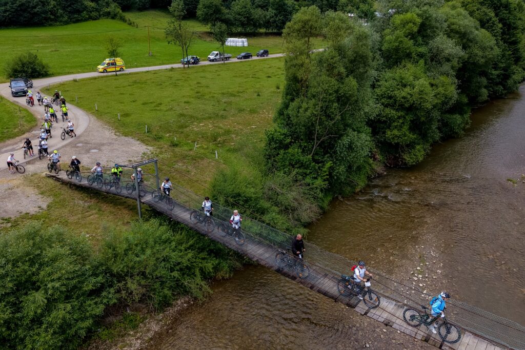 Widok z lotu ptaka na grupę rowerzystów przechodzących z rowerami przez wiszący most nad rzeką, w tle karetka pogotowia na drodze.