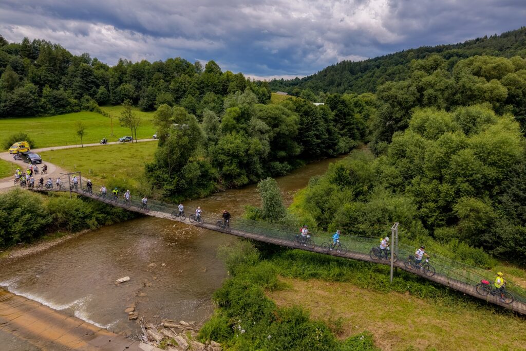 Widok z lotu ptaka na liczną grupę rowerzystów przekraczających wiszący most nad rzeką pod pochmurnym niebem.