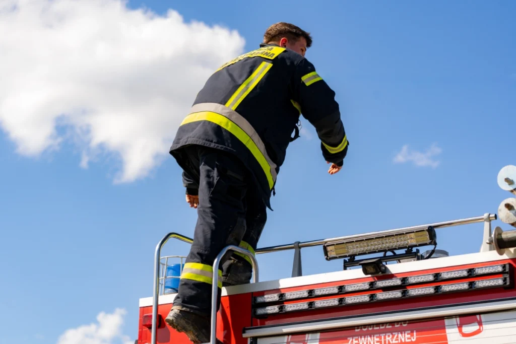A firefighter climbs a ladder onto the roof of a fire engine against a blue sky