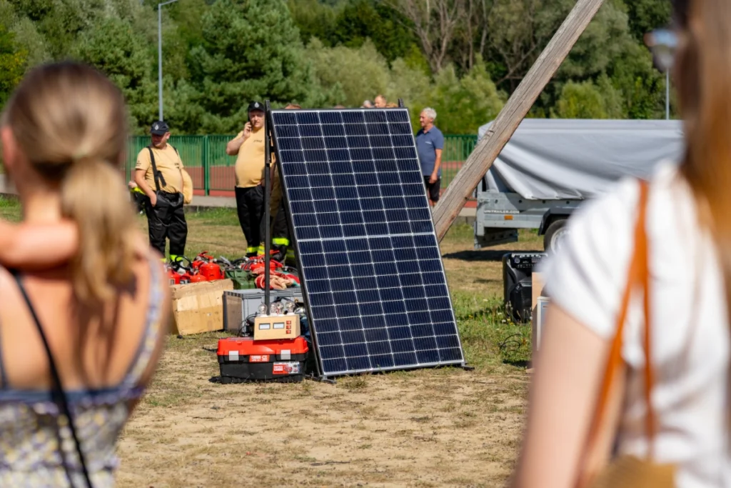 Spectators view a solar panel on the grass, behind which volunteer firefighters stand by red fire extinguishers
