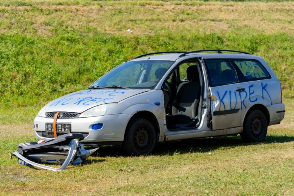 A silver car marked AUTO KUREK without its driver's door, the cut-off door lying on the grass