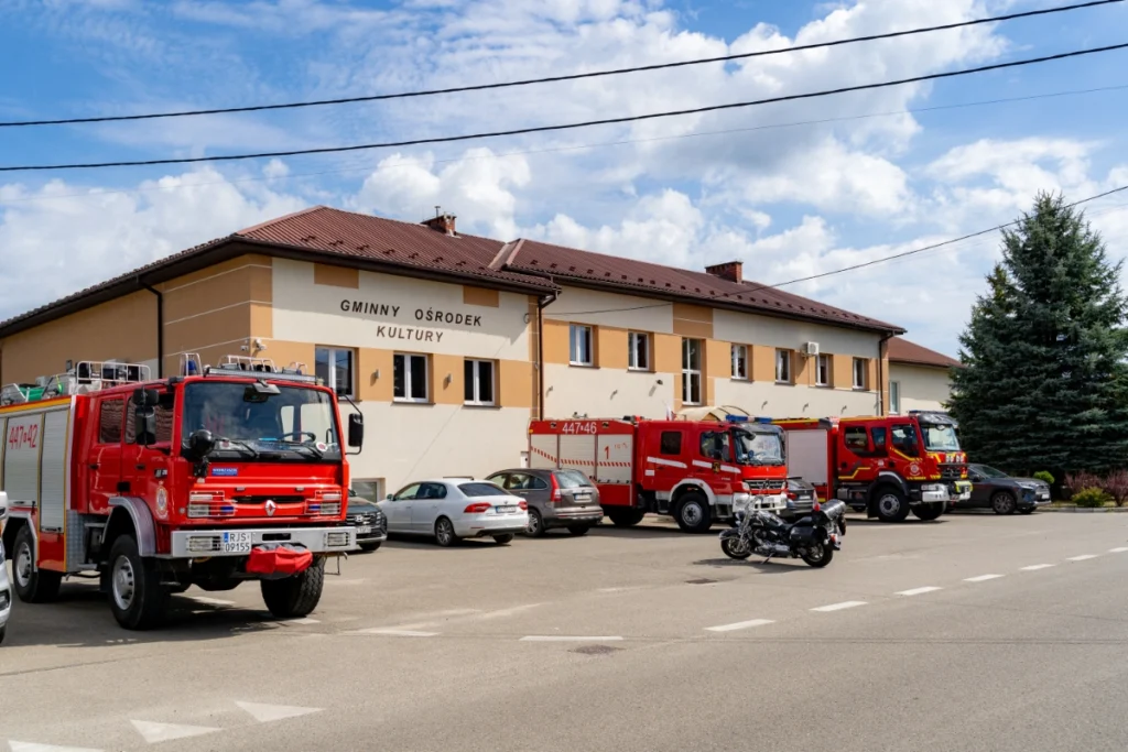 Fire engines, cars and a motorcycle parked in front of a building signed GMINNY OŚRODEK KULTURY (Municipal Cultural Centre)